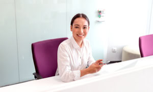 Eine Person sitzt an einem weißen Schreibtisch, trägt ein weißes Hemd und hält ein Smartphone. Im Hintergrund sind lila Stühle und eine weiße Wand zu sehen.