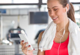 Frau im Fitnessstudio mit Handtuch um den Hals, hält ein Smartphone und trägt Kopfhörer.