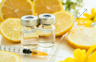 Zwei Glasfläschchen mit Flüssigkeit, eine Spritze und aufgeschnittene Zitronen auf einem Tisch, umgeben von gelben Blumen.