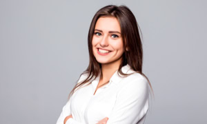 Frau mit langen braunen Haaren lächelt und trägt ein weißes Hemd vor grauem Hintergrund.