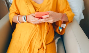 Person in gelbem Gewand sitzt auf einem Stuhl und hält ein rotes Smartphone in den Händen. Am Arm sind Krankenhausarmbänder und ein intravenöser Zugang sichtbar.