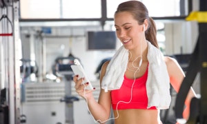 Frau im Fitnessstudio mit Handtuch um den Hals, hält ein Smartphone und trägt Ohrhörer.