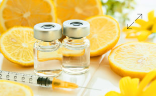 Zwei Glasfläschchen mit Flüssigkeit, eine Spritze und aufgeschnittene Zitronen auf einem Tisch mit gelben Blumen im Hintergrund.