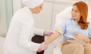 Eine Frau in einem weißen Kittel und rosa Handschuhen nimmt einer anderen Frau Blut ab, die auf einem Stuhl sitzt.