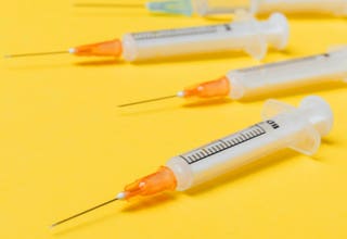 Vier Spritzen mit orangefarbenen Kappen liegen auf einem gelben Hintergrund.