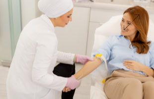 Eine Frau in einem weißen Kittel und rosa Handschuhen nimmt einer anderen Frau Blut ab, die auf einem Stuhl sitzt.