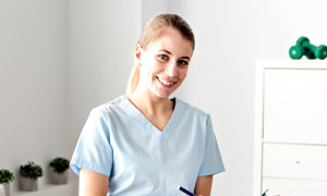 Eine Person in hellblauer medizinischer Kleidung hält einen Klemmbrett und lächelt in einem hellen Raum. Im Hintergrund sind Pflanzen und ein Regal zu sehen.