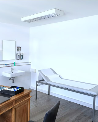 luftiges, grosszuegiges arztzimmer mit waerme ausstrahlnendem massivholzsekreter, untersuchungsliege und hygieneecke mit waschbecken und seifenspendern