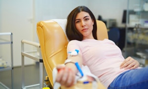 Eine Frau sitzt in einem gelben Stuhl und spendet Blut, mit einem Schlauch am Arm befestigt.