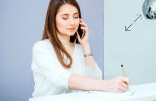 Eine Frau mit langen braunen Haaren telefoniert und schreibt gleichzeitig etwas auf ein Blatt Papier. Sie trägt ein weißes Oberteil.