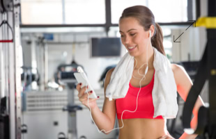 Frau im Fitnessstudio mit Handtuch um den Hals, hält ein Smartphone und trägt Kopfhörer.