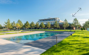 Ein rechteckiger Pool in einem gepflegten Garten mit grünen Rasenflächen und Bäumen, im Hintergrund ein großes Gebäude unter blauem Himmel.