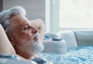 Älterer Mann mit grauem Bart entspannt sich in einem Whirlpool, mit geschlossenen Augen und den Armen hinter dem Kopf verschränkt.