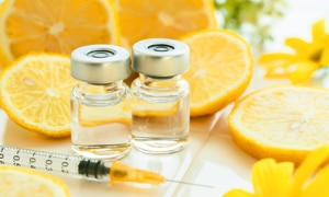 Zwei Glasfläschchen mit Flüssigkeit, eine Spritze und geschnittene Zitronen auf einem Tisch mit gelben Blumen im Hintergrund.