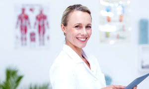 Frau in weißem Kittel hält ein Tablet in einem hellen Raum mit anatomischen Postern im Hintergrund.