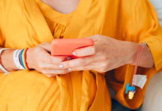 Person in einem gelben Kleid sitzt auf einem Stuhl und hält ein rotes Smartphone in den Händen. Am Arm sind mehrere Armbänder und ein medizinischer Zugang zu sehen.