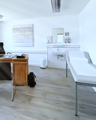 grosszuegiges und luftig wirkendes arztzimmer mit waerme ausstrahlnendem massivholzsekreter im zentrum, untersuchungsliege und hygieneecke mit waschbecken und seifenspendern