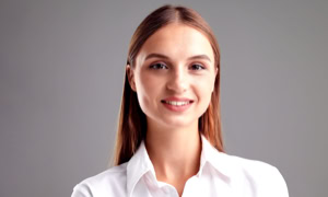 Eine lächelnde Person mit langen braunen Haaren trägt ein weißes Hemd vor einem grauen Hintergrund.