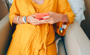 Person in gelbem Kleid sitzt auf einem Stuhl und hält ein rotes Smartphone, mit Krankenhausarmbändern am Handgelenk.
