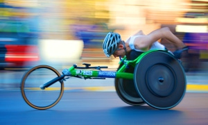 Person in einem grünen Rennrollstuhl, die einen Helm trägt und sich schnell bewegt, mit verschwommenem Hintergrund.