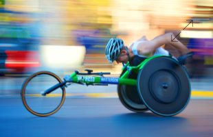 Person in einem grünen Rennrollstuhl, die einen Helm trägt und sich schnell bewegt, mit verschwommenem Hintergrund.