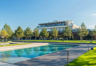 Ein rechteckiger Pool in einem Park mit Bäumen und einem großen Gebäude im Hintergrund unter blauem Himmel.
